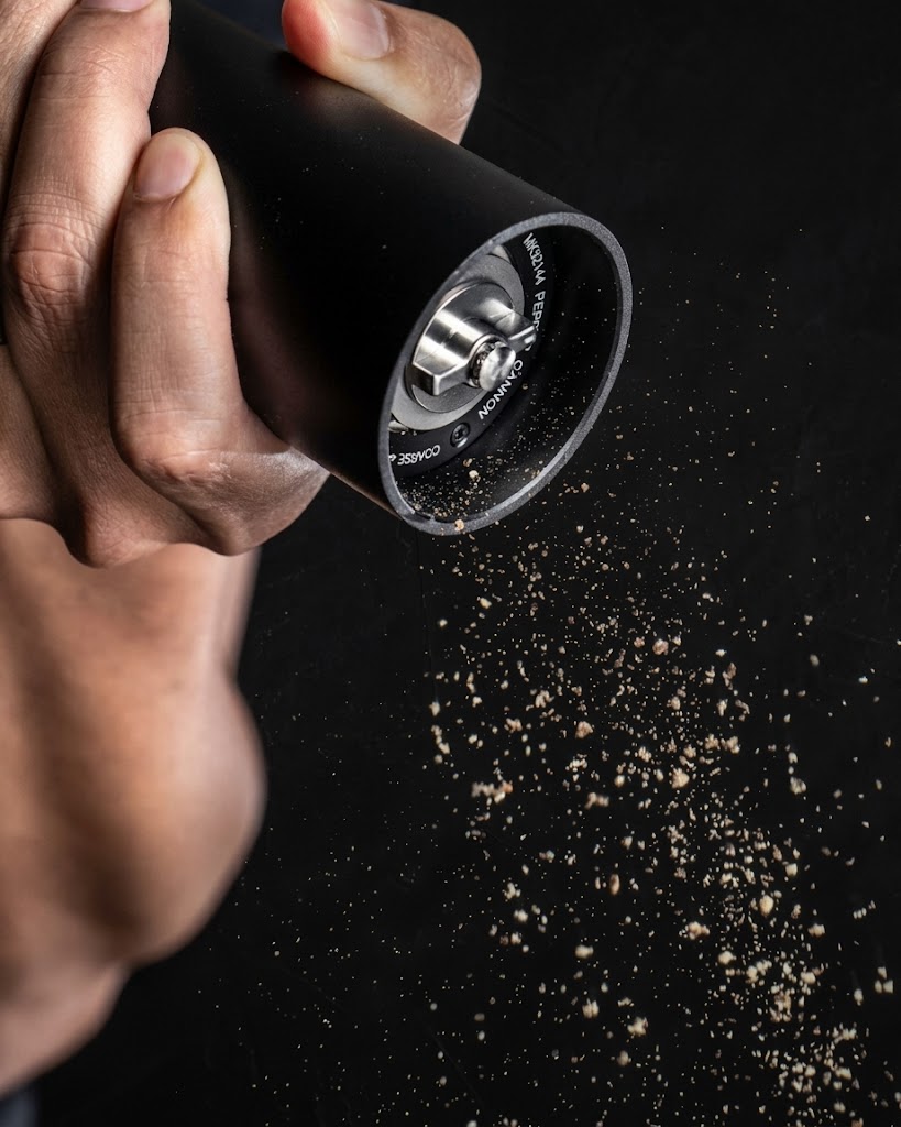 A hand grinding fresh pepper from the Pepper Cannon onto a dark surface.