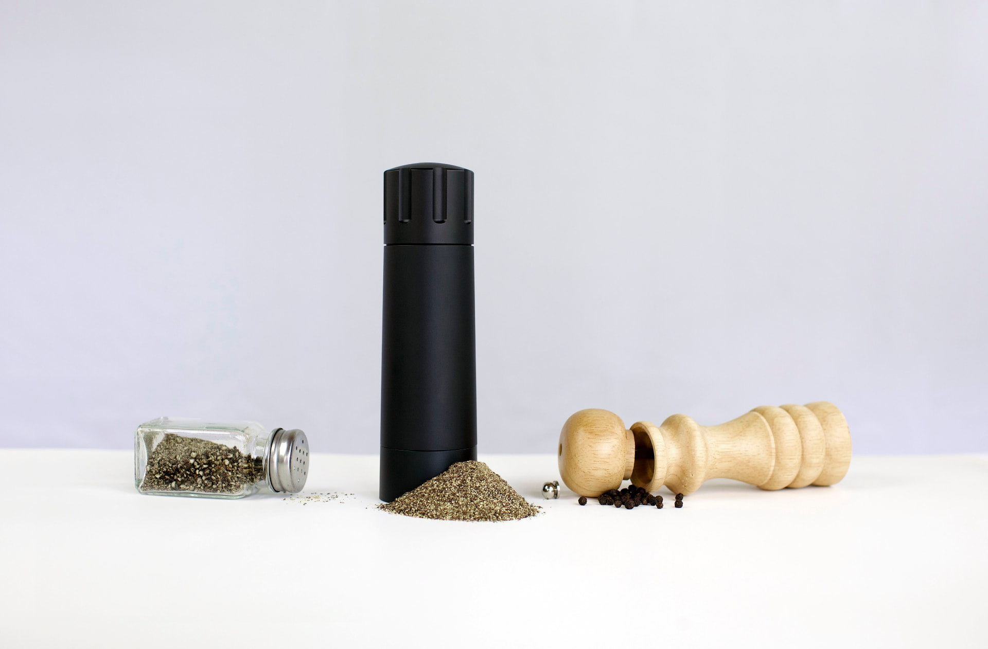 The Pepper Cannon between a shaker of pre-ground pepper and a wooden mill, with a mound of fresh pepper.