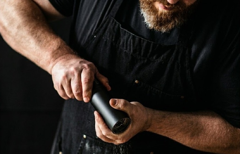A bearded man in a black apron grinding pepper with the Pepper Cannon.